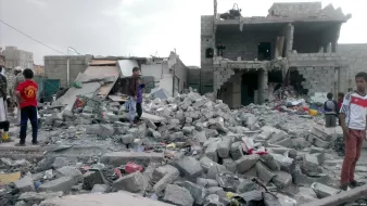 L'image montre des bâtiments gravement endommagés, avec des débris éparpillés sur le sol. On peut voir des enfants et des adultes présents parmi les ruines, cherchant peut-être des effets personnels ou évaluant les dégâts. L'environnement semble chaotique et témoigne d'une destruction significative, probablement suite à un conflit ou à une catastrophe. Le ciel est nuageux, ce qui ajoute à l'atmosphère sombre de la scène.