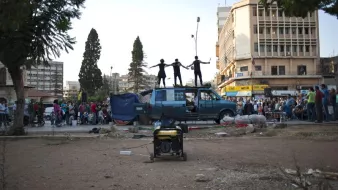 L'image montre une scène animée dans un endroit urbain. Au premier plan, on voit un espace dégagé avec un générateur. En arrière-plan, un certain nombre de personnes sont rassemblées, probablement pour assister à un événement. Sur le toit d'une fourgonnette bleue, trois personnes sont perchées, probablement en train de performer ou de s'exprimer de manière artistique. L'environnement semble être un mélange de vie urbaine et de rassemblement communautaire.