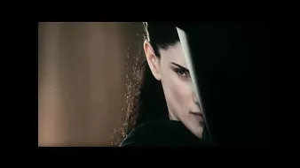 L'image montre une femme avec une expression intense et déterminée. Elle est à moitié cachée derrière un objet, ce qui accentue son regard perçant. Ses cheveux sont longs et foncés, et son attitude suggère qu'elle est dans une situation de confrontation ou de défense. L'éclairage met en avant son visage et crée une ambiance mystérieuse.