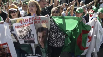L'image montre une manifestation où des personnes sont rassemblées, exprimant des revendications. On peut voir des individus brandissant des pancartes et des drapeaux, notamment un drapeau algérien. Les manifestants semblent mobilisés et engagés, scandant des slogans pour la libération de Mustapha Aouissi. L'atmosphère semble dynamique et pleine d'énergie, avec une forte présence de diversité parmi les participants.