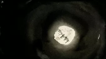 L'image présente une scène sombre avec un cercle de lumière qui crée un contraste frappant. Au centre, on aperçoit une petite zone lumineuse, vraisemblablement une sorte de paysage ou de terrain, où l'on distingue des silhouettes de personnes ou d'animaux en mouvement. Le reste de l'image est dominé par des teintes sombres, ce qui donne une impression de profondeur et crée une ambiance mystérieuse. Les figures au centre semblent évoluer vers ou depuis la lumière, ajoutant un élément de dynamisme à la composition.