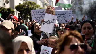 L'image montre une foule rassemblée pour une manifestation. Au premier plan, une femme porte un enfant sur ses épaules, et elle tient un panneau avec le mot "DEGAGE". Les manifestants semblent exprimer des revendications politiques. On peut également voir d'autres panneaux en arabe et des drapeaux, ce qui indique une atmosphère de protestation collective. La scène se déroule en plein air, probablement dans une ville, et les expressions des participants montrent une forte détermination.
