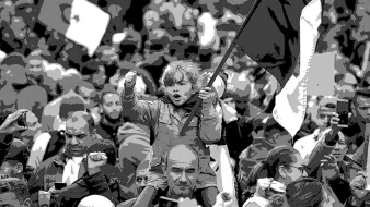L'image montre une foule rassemblée lors d'une manifestation. Au premier plan, une petite fille se tient sur les épaules d'un adulte, brandissant un drapeau. Elle semble enthousiaste et déterminée, levant le poing. La foule autour d'elle est dense, avec des visages variés et des drapeaux visibles en arrière-plan. L'atmosphère est dynamique et engagée, évoquant un sentiment collectif et de solidarité.