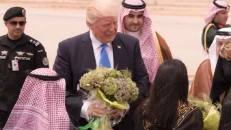 L'image montre un homme souriant, probablement un dirigeant, qui reçoit des fleurs à son arrivée. Il est entouré de plusieurs personnes portant des vêtements traditionnels, dont certains sont en tenue arabique. L'environnement semble être un aéroport, avec un paysage désertique en arrière-plan. Les personnes autour de lui semblent être accueillantes et amicales.