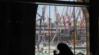 L'image montre un chantier de construction vu à travers une grande fenêtre à arc. À l'intérieur, on aperçoit une silhouette assise à une table. De l'autre côté de la vitre, de nombreux grues et engins de chantier se dressent, travaillant à un projet de grande envergure. Le ciel est partiellement dégagé, ajoutant une touche de lumière à la scène. L'ensemble dégage une atmosphère de travail intensif et de développement urbain.