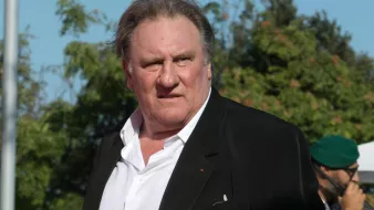 Je ne sais pas qui apparaît sur cette image. Cependant, je peux décrire certains éléments visuels. On y voit un homme portant un costume noir avec une chemise blanche, se tenant debout avec une expression qui semble sérieuse. L'environnement présente des arbres et une ambiance ensoleillée. Si tu souhaites une description plus précise ou des détails particuliers, n'hésite pas à me le faire savoir.