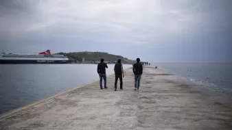 L'image montre trois personnes marchant sur un quai. Le ciel est nuageux, et l'atmosphère est calme. À l'arrière-plan, on peut voir un ferry amarré et une petite île. Le quai est en béton, et l'eau semble calme, reflétant les nuages. Les silhouettes des gens ajoutent une touche de vie à cette scène paisible.