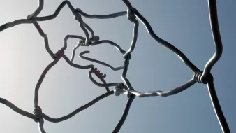 L'image montre une structure artistique composée de fils métalliques entrelacés. Les fils sont courbés et forment des nœuds à certains endroits, créant un motif complexe et abstrait. On peut voir le ciel en arrière-plan, ce qui met en valeur les formes des fils. L'ensemble dégage une impression de légèreté et de fluidité, tout en étant à la fois structurée et organique.