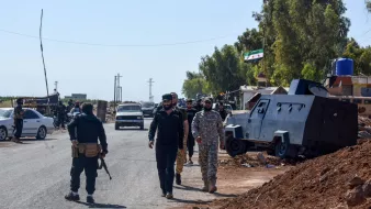 Des soldats marchent le long d'une route, près de véhicules militaires et d'arbres.