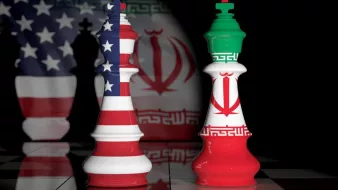 L'image représente des pièces d'échecs en forme de rois, une avec les couleurs du drapeau américain et l'autre avec celles du drapeau iranien. En arrière-plan, on peut voir des ombres ou des motifs représentant ces deux pays. La scène évoque une confrontation symbolique entre les États-Unis et l'Iran, souvent associée à des tensions géopolitiques. Le fond sombre renforce l'aspect dramatique de l'image.