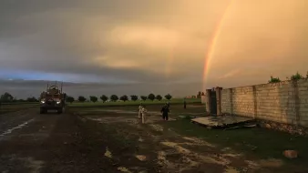 L'image montre un paysage rural sous un ciel dramatique, avec des nuages sombres et un ciel éclairci. On peut voir un arc-en-ciel double qui s'étend dans le ciel, ajoutant une touche de couleur. À gauche, un véhicule militaire est garé sur un chemin boueux, tandis qu'à droite, deux personnes marchent près d'un mur en béton. Le sol est humide, témoignant d'une récente pluie, et des arbres sont visibles en arrière-plan, soulignant une atmosphère paisible malgré la présence militaire.