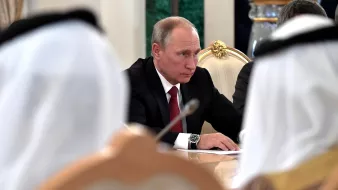 L'image montre un homme assis à une table lors d'une réunion. Il porte un costume noir avec une cravate rouge et semble concentré sur la discussion. Autour de lui, on peut voir des personnes en vêtements traditionnels, comme des robes blanches et des turbans. L'environnement est formel, avec des décorations élégantes. L'atmosphère paraît sérieuse et impliquée dans des délibérations.