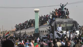 L'image montre une grande foule de personnes rassemblées autour et sur un train. Beaucoup de gens se tiennent sur le toit du train, tandis que d'autres s'accrochent aux côtés. L'atmosphère semble vibrante et énergique, probablement liée à une manifestation ou un événement important. En arrière-plan, on peut apercevoir des bâtiments et des infrastructures typiques d'une ville. La scène dégage un sentiment de solidarité et de détermination parmi les participants.