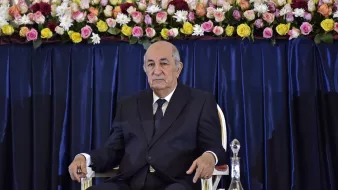 L'image montre un homme assis sur une chaise, portant un costume sombre et une cravate. Il a l'air sérieux et attentif. À l'arrière-plan, il y a une décoration florale composée de roses et d'autres fleurs qui ajoutent de la couleur à la scène. La disposition des fleurs est soignée, suggérant un cadre formel ou officiel.