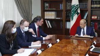 L'image montre une réunion dans un bureau. Un homme, probablement un leader politique, est assis à droite, face à une table en bois. Il semble concentré sur des documents. À sa gauche, il y a trois personnes, vraisemblablement des conseillers ou des diplomates, qui prennent des notes et écoutent attentivement. Tous portent des masques. En arrière-plan, on voit une bibliothèque remplie de livres et le drapeau du Liban. L'atmosphère semble formelle et sérieuse.