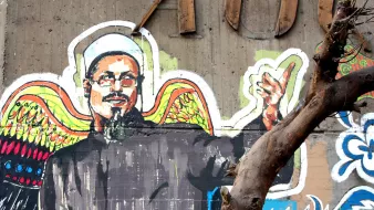 L'image représente une fresque murale colorée. On y voit un homme aux épaules larges, vêtu d'une tunique noire et d'un chapeau traditionnel. Il a un regard expressif et semble faire un geste de la main, comme s'il s'adressait à quelqu'un. En arrière-plan, des motifs vibrants et contrastés, dont des ailes colorées, ajoutent une dimension artistique à la composition. Un arbre, avec des branches dénudées, est également présent au premier plan, soulignant l'environnement urbain de la fresque.