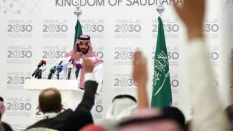 L'image montre une conférence de presse organisée en Arabie Saoudite. Un homme, vêtu d'une robe traditionnelle saoudienne et d'un headdress, se tient devant un podium avec plusieurs microphones. En arrière-plan, on peut voir des drapeaux saoudiens ainsi qu'un panneau affichant des informations sur la Vision 2030 de l'Arabie Saoudite. Plusieurs personnes sont présentes, levant les mains, probablement pour poser des questions. L'atmosphère semble sérieuse, centrée sur des discussions importantes liées à l'avenir du pays.