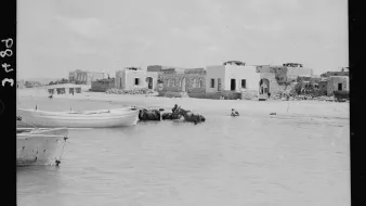 L'image montre une scène maritime où plusieurs bateaux, probablement des barques, sont amarrés au bord d'une eau peu profonde. En arrière-plan, on peut apercevoir des bâtiments en ruines, suggérant une ancienne architecture. À proximité, un homme semble se trouver dans l'eau, tandis qu'un autre est à cheval ou monté sur un animal aquatique. Le ciel est nuageux, apportant une lumière diffuse à l'ensemble de la scène. L'atmosphère semble tranquille et un peu nostalgique, évoquant un passé révolu.