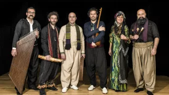 L'image montre un groupe de six musiciens posant ensemble sur scène. Ils sont vêtus de vêtements traditionnels, ce qui évoque une ambiance culturelle riche. Chaque membre tient un instrument différent : on peut voir un instrument à cordes, des percussions et d'autres instruments typiques. Leur expression est joyeuse et conviviale, et l'éclairage met en valeur leurs costumes. L'arrière-plan est sombre, ce qui fait ressortir leurs tenues colorées et détaillées. Cela donne une impression de performance musicale authentique et festive.