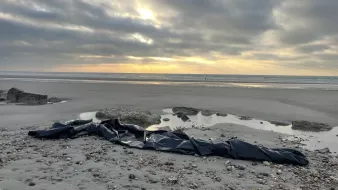 L'image montre une plage désertique au crépuscule, avec un ciel nuageux où quelques rayons de soleil percent à travers les nuages. Sur le sable, on aperçoit un long objet noir qui semble être un morceau de toile ou de bâche, posé près de quelques rochers. À l'horizon, la mer et quelques petites vagues reflètent la lumière tamisée du ciel. L'atmosphère est à la fois calme et mystérieuse.