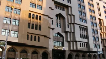 L'image montre un bâtiment architectural impressionnant, avec une façade en pierre et de grandes fenêtres ornées. Les éléments décoratifs évoquent un style inspiré de l'architecture islamique, notamment avec des arches et des motifs géométriques distincts. La lumière du soleil met en valeur les détails de la structure, tandis que des lampadaires et des lampes décoratives sont visibles au premier plan. Cela dégage une ambiance à la fois moderne et traditionnelle.