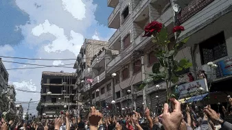 L'image montre une grande foule au milieu d'une rue, probablement lors d'une manifestation. Les gens semblent levés les bras, tenant des roses, ce qui suggère une ambiance pacifique ou festive. En arrière-plan, on aperçoit des bâtiments et un ciel partiellement nuageux. L'atmosphère semble être chargée d'émotions et d'unité entre les participants.