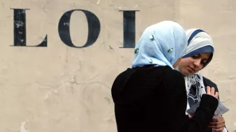 L'image présente deux femmes discutant, l'une d'elles portant un hijab bleu clair et l'autre un hijab sombre. Elles semblent concentrées sur un document ou une feuille qu'elles regardent ensemble. En arrière-plan, on peut voir le mot "LOI" écrit en lettres majuscules sur un mur beige, ce qui donne un contexte à la scène. L'atmosphère semble calme et introspective.