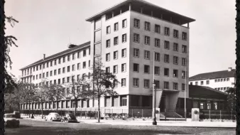 L'image représente un bâtiment architectural de plusieurs étages, probablement de style moderne. Il est photographié sous une lumière naturelle, mettant en avant sa façade aux lignes géométriques. Le bâtiment semble être entouré d'arbres et de quelques véhicules anciens, ce qui donne une ambiance d'époque. On peut également apercevoir des éléments de la rue, comme des lampadaires et éventuellement d'autres bâtiments en arrière-plan. L'ensemble de l'image présente une esthétique monochrome, accentuant le caractère historique de la scène.