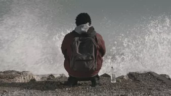 L'image montre une personne assise sur un rivage, tournant le dos à la caméra. Elle porte une veste et un sac à dos. À côté d'elle, il y a une bouteille d'eau. On peut voir des vagues qui se brisent sur les rochers, créant un effet de mouvement dans l'eau. L'atmosphère semble calme mais dynamique en raison du mouvement des vagues.
