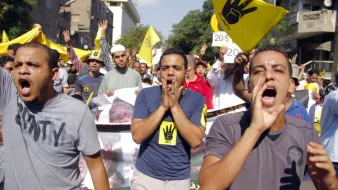 L'image montre une manifestation animée, avec des participants portant des t-shirts colorés. Ils expriment des slogans, certains scandent en levant les bras, tandis que d'autres tiennent des pancartes, notamment avec un symbole en forme de main. L'ambiance semble être pleine d'énergie et de détermination, reflétant une forte mobilisation autour d'une cause. Les bâtiments en arrière-plan suggèrent un milieu urbain.