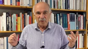 L'image montre un homme debout devant une étagère remplie de livres. Il porte une chemise à rayures et semble s'adresser à un public ou donner une présentation. Ses mains sont levées, ce qui indique qu'il est en train de faire un point ou d'expliquer quelque chose. L'arrière-plan est constitué de livres aux couvertures variées, suggérant un environnement intellectuel ou académique.