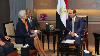 L'image montre une réunion entre des personnalités importantes. On peut voir une femme en tenue professionnelle, accompagnée d'un homme qui semble prendre des notes ou écouter attentivement. Ils sont assis en face d'un homme, probablement en position d'autorité, qui est vêtu d'un costume. En arrière-plan, il y a un drapeau égyptien et quelques éléments de décoration, comme des fleurs. L'ambiance semble formelle et axée sur la discussion.