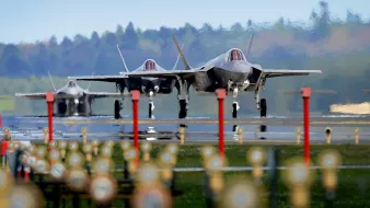 L'image montre plusieurs avions de chasse F-35 alignés sur une piste d'atterrissage. Au premier plan, on peut voir des lumières et des repères au sol, tandis que les avions sont prêts au décollage. En arrière-plan, on aperçoit une forêt verdoyante sous un ciel clair. L'ambiance général semble être celle d'une base aérienne, mettant en avant la puissance et la technologie militaire moderne.