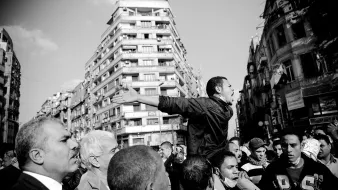 L'image montre une scène de manifestation dans un environnement urbain. Au centre, un homme se tient debout, les bras écartés, semblant exprimer une idée forte ou appeler à l'action. Il est entouré d'une foule dense, composée d'hommes, qui l'écoute ou réagit à son discours. Les bâtiments en arrière-plan montrent une architecture typique de ville, et l'image est en noir et blanc, ce qui accentue les émotions et la tension de la situation. L'atmosphère semble dynamique et engageante.