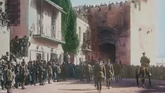 Cette image, en couleur, montre une scène historique se déroulant probablement lors d'un événement important à Jérusalem. Au premier plan, des soldats en uniforme, dont certains à cheval, avancent avec une certaine autorité. En arrière-plan, il y a une porte majestueuse sur un mur ancien, entourée de personnes qui regardent la procession depuis des balcons et des hauteurs. La foule est dense, avec des hommes, des femmes et des enfants, tous attentifs à ce qui se passe. Les bâtiments environnants sont typiques de l'architecture de la région, et l'atmosphère semble être à la fois festive et solennelle.
