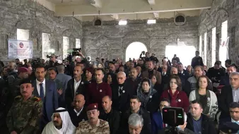 L'image montre une grande foule de personnes rassemblées dans un bâtiment à l'apparence industrielle ou historique, avec des murs en pierre. Les participants semblent être assis ou debout et portent des badges ou des pin's. Diverses personnes de différentes origines apparaissent, y compris des représentants militaires en uniforme. L'atmosphère semble formelle, suggérant un événement significatif ou une réunion. Des caméras et des journalistes sont également présents, indiquant qu'il pourrait s'agir d'une conférence ou d'une célébration.