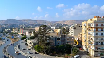 L'image montre un paysage urbain avec des bâtiments variés, y compris des immeubles résidentiels et commerciaux, ainsi qu'une route serpentine. Au loin, on peut voir des collines et un ciel clair avec quelques nuages. La scène dégage une ambiance paisible, avec des voitures circulant sur la route et des arbres ajoutant une touche de verdure à l'environnement.