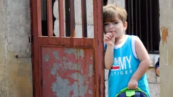 L'image montre un jeune garçon se tenant dans l'encadrement d'une porte. Il porte un débardeur bleu avec des inscriptions et tient un sac à dos coloré. Le garçon semble pensif, en train de sucer son pouce. La porte derrière lui est rustique et en métal, avec des jeux d'ombre et de lumière. L'environnement semble simple et authentique, suggérant une ambiance tranquille.