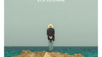 L'image représente une affiche de film intitulée "It Must Be Heaven", réalisé par Elia Suleiman. On y voit un homme de dos, portant un chapeau, se tenant debout sur un rocher au bord de la mer. L'horizon est dégagé, avec un ciel bleu clair. Le titre du film est inscrit en lettres blanches en bas de l'affiche, avec également le nom du réalisateur. L'ensemble de l'image évoque une ambiance contemplative et poétique.