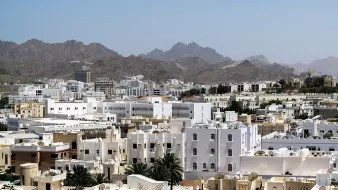 L'image montre une vue panoramique d'une ville située dans une région montagneuse. On peut voir de nombreux bâtiments aux façades blanches et crème, qui typiquement se distinguent dans un environnement aride. Les montagnes en arrière-plan ajoutent une dimension naturelle impressionnante au paysage urbain. Le ciel est dégagé et ensoleillé, accentuant la lumière qui se reflète sur les bâtiments. Cette scène évoque une atmosphère calme et sereine.