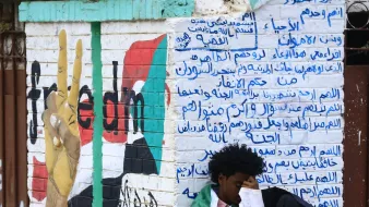 L'image montre un mur peint avec un symbole de paix, probablement représentant un mouvement pour la liberté. À gauche, il y a un graffiti d'une main levant deux doigts, symbolisant le mouvement pacifiste. Sur le mur, des inscriptions en arabe sont visibles, évoquant probablement des messages politiques ou sociaux. Au premier plan, un jeune homme est assis, enroulé dans un drapeau, semblant pensif ou mélancolique. L'atmosphère suggère un moment de réflexion sur des luttes pour la liberté.