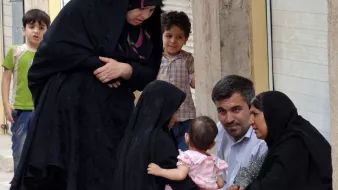 L'image montre un groupe de personnes dans une rue. Deux femmes, vêtues de vêtements noirs, sont assises sur le sol et discutent. L'une d'elles tient un bébé, qui porte une légère robe rose. À proximité, un homme regarde les femmes et semble participer à la conversation. Un jeune garçon, en arrière-plan, observe la scène. L'environnement apparaît urbain, avec des bâtiments en briques visibles. L'atmosphère semble conviviale et familiale.