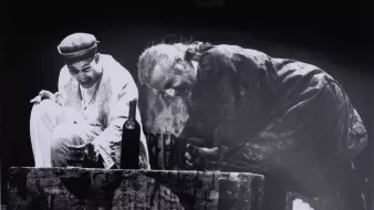 Deux hommes en scène, riant, près d'une bouteille sur une caisse au fond sombre.