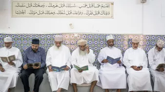 L'image montre un groupe d'hommes assis en ligne, lisant des livres, probablement des copies du Coran. Ils portent des vêtements traditionnels, tels que des dishdashas et des couvre-chefs. En arrière-plan, on peut voir un mur décoré de carrelage coloré, sur lequel est inscrite une citation ou un texte. L'atmosphère semble calme et sereine, suggérant un moment de prière ou de méditation.