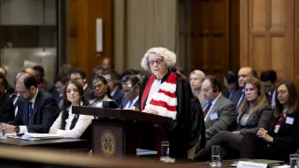 L'image montre une scène dans une salle d'audience, probablement dans un tribunal. Une femme, vêtue d'une robe magistrale et d'une écharpe, se tient derrière un pupitre en train de prendre la parole. Plusieurs personnes sont assises à des tables devant elle, écoutant attentivement. L'environnement semble formel, avec des éléments architecturaux en bois et des fenêtres qui apportent de la lumière naturelle. L'atmosphère est solennelle et concentrée, indicative d'une procédure judiciaire importante.