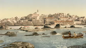 L'image représente un paysage côtier avec une ville en arrière-plan. On peut voir des maisons blanches construisant un front de mer, accompagnées d'une église au sommet d'une colline. Au premier plan, deux personnes naviguent dans un petit bateau sur une eau calme, entouré de rochers. Le ciel est dégagé, suggérant une journée ensoleillée, et la scène évoque une atmosphère paisible et pittoresque. Les couleurs sont vives, mettant en valeur la beauté naturelle de l'endroit.