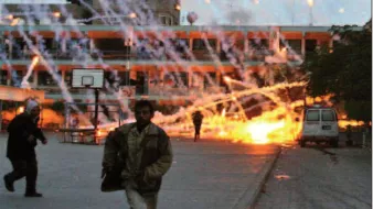 L'image montre une scène de chaos avec des explosions et des traînées de lumière dans le ciel. Des personnes semblent fuir dans une zone qui ressemble à une cour d'école, tandis qu'un bâtiment en arrière-plan est visible. L'environnement semble marqué par une tension intense, avec des traces d'incendie et des éclats lumineux qui suggèrent une situation dangereuse. L'atmosphère est sombre, renforçant le sentiment d'urgence et de désarroi.