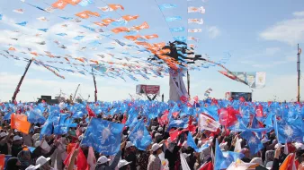L'image montre une grande foule rassemblée lors d'un événement, probablement un rassemblement politique ou une manifestation. Des drapeaux de différentes couleurs, notamment bleu et orange, flottent dans l'air, créant une ambiance festive. Les participants portent des chapeaux blancs et semblent très engagés dans l'événement. Le ciel est clair, ce qui ajoute à l'énergie de la scène. En arrière-plan, des affiches et des structures indiquent un cadre organisé pour cet événement public.