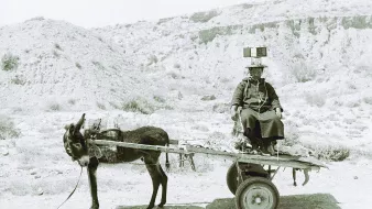 L'image montre une scène en noir et blanc d'un homme assis sur un chariot tiré par un âne. L'homme porte des vêtements traditionnels et semble se diriger quelque part, tandis que l'âne est attaché à l'avant du chariot. L'arrière-plan présente un paysage aride et désertique, avec des collines ou des montagnes à l'horizon. L'atmosphère de la photographie évoque un sentiment de simplicité et de vie rurale.