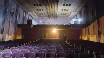L'image montre l'intérieur d'un théâtre ancien. On peut voir des rangées de sièges en velours, qui semblent un peu usés, dirigées vers une scène surélevée. La scène est éclairée par une lumière vive, tandis que le reste de la salle est dans une ambiance plus sombre. On aperçoit également des détails architecturaux au plafond et sur les murs, qui évoquent un style vintage. Un homme se tient debout au fond, ajoutant une touche humaine à cet espace, qui dégage une atmosphère à la fois nostalgique et mystérieuse.